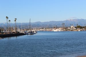 Calm morning view of Channel Islands Harbor in Oxnard, California, featuring sailboats, palm trees, and distant mountains reflected on the still water.