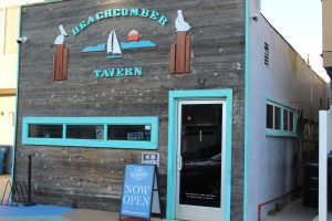 Exterior of the Beachcomber Tavern with weathered wood siding, turquoise trim, nautical signage, and a “Now Open” sign at the entrance.