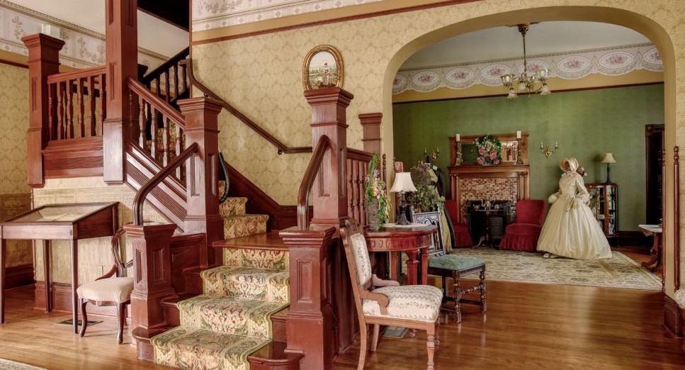 “Interior of the Camarillo Ranch House featuring a carved wooden staircase, Victorian furnishings, and period décor.”