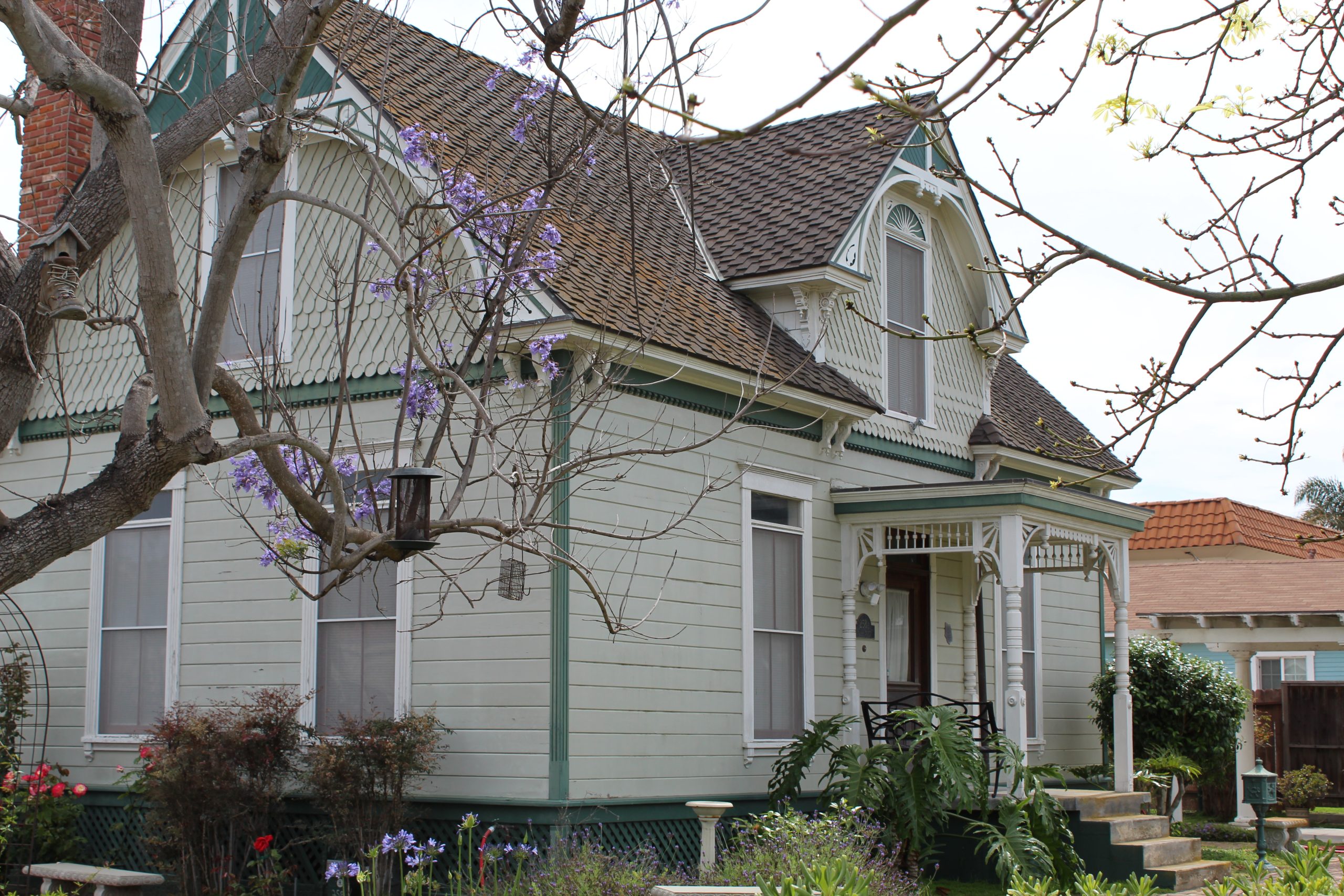 “Vintage cottage-style home with steep roof, decorative trim, pastel siding, blooming garden, and purple-flowering tree in a quiet Port Hueneme neighborhood.”