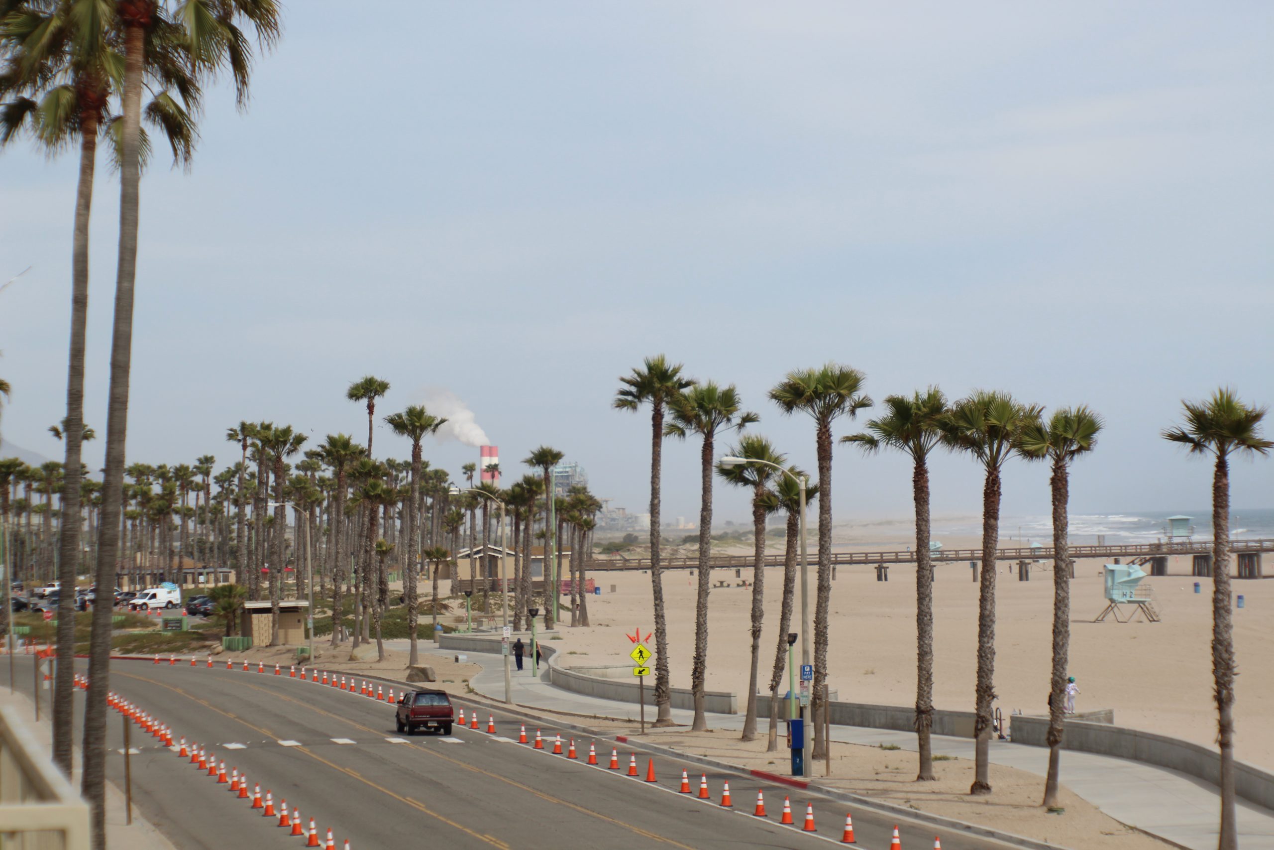 “Coastal road lined with palm trees, beach walkway, parked cars, traffic cones, and a long pier overlooking the ocean in Port Hueneme.”